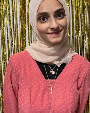 A picture of Aisha in a pink top standing in front of a gold background.